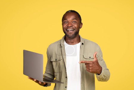 Photo for Happy mature black man in casual showing finger at computer, chatting in social networks, isolated on orange studio background. Advice for blog, gadget for communication, work remotely, ad and offer - Royalty Free Image