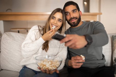 Téléchargez les photos : Cheerful young arab guy with beard and european lady watch tv with remote control, eat popcorn, enjoy video, spare time, sit in living room interior. Fun together, movie evening, love, relationship - en image libre de droit