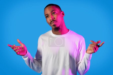 Téléchargez les photos : Confused unsure handsome african american young man in white gesturing with arms up on blue studio background, looking at copy space. Human gestures concept - en image libre de droit