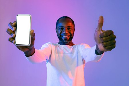 Téléchargez les photos : Smiling handsome middle aged black man in casual showing smartphone with white blank screen in his hand and thumb up, recommending mobile app, mockup, neon studio background - en image libre de droit
