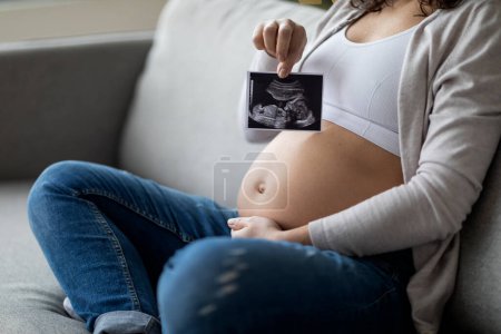 Téléchargez les photos : Femme enceinte méconnaissable montrant une photo d'échographie de bébé assis sur le canapé à la maison, femme enceinte montrant la première photo de son enfant, profitant d'une grossesse en bonne santé, recadrée - en image libre de droit