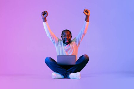 Téléchargez les photos : Emotional bearded handsome mature african american man in white with computer on lap sitting on floor, raising hands up and screaming over neon studio background, trading or gaming, copy space - en image libre de droit