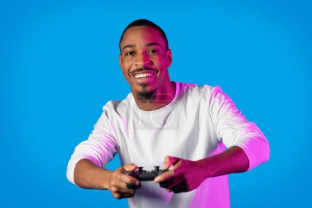 Foto de Feliz alegre sonriente joven afroamericano en blanco jugando videojuegos con joystick sobre fondo azul en luz de neón, chico negro disfrutando de su tiempo, espacio de copia - Imagen libre de derechos