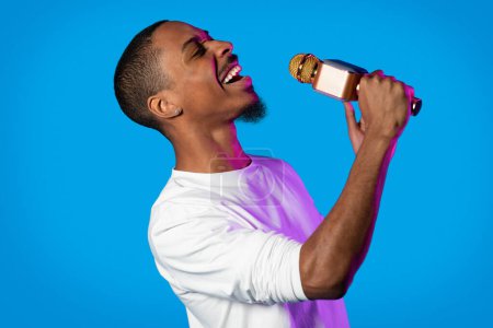 Téléchargez les photos : Talented charismatic emotional bearded young black guy in white longsleeve singing with closed eyes on blue studio background in neon light, holding microphone - en image libre de droit