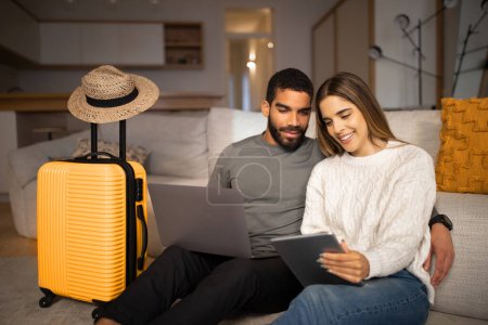 Téléchargez les photos : Smiling young arab male, european lady use tablet and laptop to choose country for vacation travel, buy tickets in living room interior with suitcase, hat. Travel app for tourists, love relationship - en image libre de droit