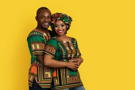 Foto de Hermosa pareja negra encantadora en trajes africanos nacionales brillantes abrazando y sonriendo a la cámara, de pie sobre el fondo del estudio amarillo, espacio de copia. Amor, matrimonio, concepto de relaciones - Imagen libre de derechos