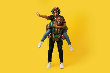 Photo for Happy beautiful black couple in traditional african costumes posing on yellow studio background, pretty young woman piggybacking her handsome middle aged husband and waving, copy space, full length - Royalty Free Image