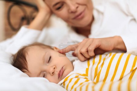 Foto de Mujer joven acariciando a su pequeña niña durmiendo linda, mujer tocando su mejilla y sonriendo, disfrutando de un momento dulce, acostados juntos en la cama, se centran en su hija - Imagen libre de derechos