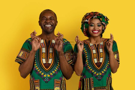 Photo for Loving attractive adult black man and woman in bright national african clothing posing on yellow studio background, holding fingers crossed with closed eyes, believe in superstitions, good luck - Royalty Free Image