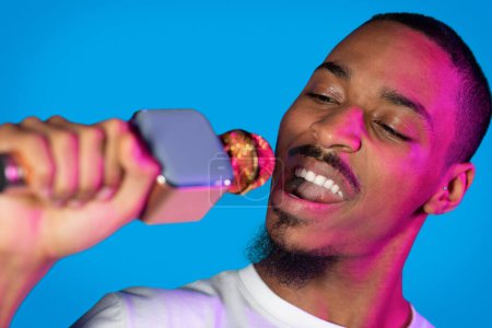 Téléchargez les photos : Cool handsome charismatic young black guy in white entertainer or singer posing with microphone in his hand over blue studio background in neon light, singing with closed eyes, closeup - en image libre de droit