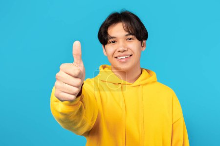 Téléchargez les photos : Portrait Of Cheerful Japanese Teen Guy Gesturing Thumbs Up Approving Great Offer Smiling To Camera Standing Posing In Studio Over Blue Background. I Like It, Approval Concept - en image libre de droit