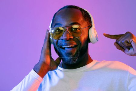 Photo for Cheerful middle aged black man wearing sunglasses using brand new wireless white headphones, pointing at gadget while listening to music over studio background, recommending nice gadget, closeup - Royalty Free Image