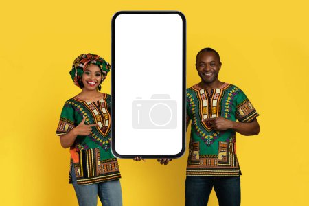 Téléchargez les photos : Positive happy bright loving black couple in traditional african costumes holding big phone together, pointing at white blank screen and smiling, showing online offer, yellow background, mockup - en image libre de droit