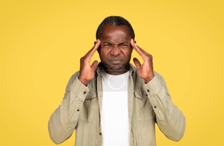 Photo for Sad adult black man in casual presses his hands to temples, suffers from headache and migraine, isolated on yellow background, studio. Facial expression, emotions, health problems, stress, pressure - Royalty Free Image