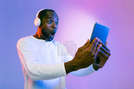 Téléchargez les photos : Shocked emotional mature black man using wireless headphones and modern digital tablet on neon studio background, looking at shining pad screen and grimacing, collage - en image libre de droit