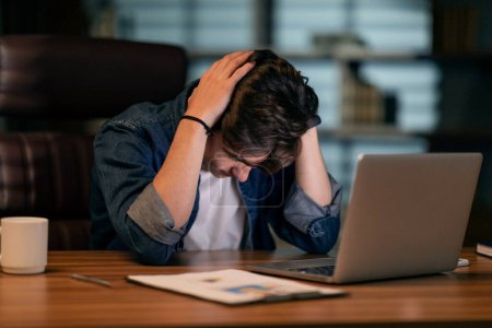 Photo for Burnout at work, overworking concept. Exhausted young man sitting at workplace in front of computer at dark office, touching his head, lack of ideas, looking for creative business solution, copy space - Royalty Free Image