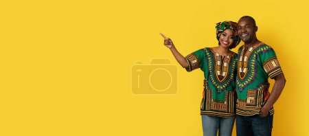 Photo for Positive cheerful african american lovers wearing colorful traditional costumes embracing and showing copy space for advertisement, black couple posing on yellow studio background, web-banner - Royalty Free Image