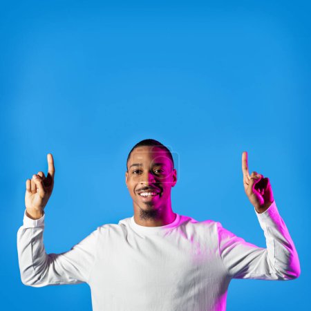 Téléchargez les photos : Positif joyeux beau jeune homme noir décontracté pointant vers le haut à l'espace de copie pour la publicité ci-dessus, montrant une offre ou une affaire agréable, posant sur la lumière au néon sur fond de studio bleu - en image libre de droit