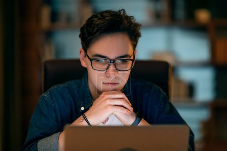 Photo for Focused IT worker young handsome guy wearing eyeglasses looking at laptop screen, employee working late at office, closeup shot, copy space. Career, job, burnout concept - Royalty Free Image