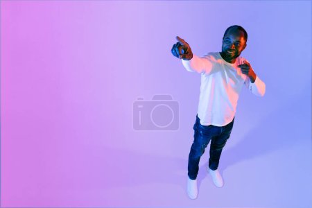 Téléchargez les photos : Handsome middle aged african american man pointing at free space for advertisement or text and smiling over colorful neon studio background, high angle view shot, full length - en image libre de droit
