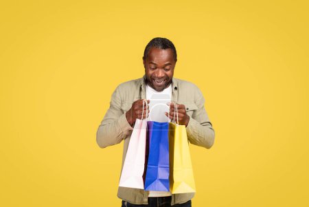 Téléchargez les photos : Glad shocked middle aged black male shopaholic in casual looking in package with purchases, enjoy fashion isolated on yellow background, studio. Huge sale, shopping, gift buying, new clothes emotions - en image libre de droit
