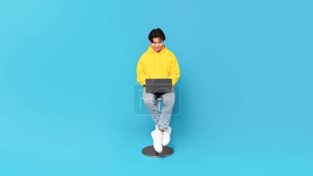 Photo for Korean Teen Guy Using Laptop Browsing Internet And Doing Homework Online On Computer Sitting Posing On Chair Over Blue Studio Background. E-Learning And Technology Concept. Panorama - Royalty Free Image