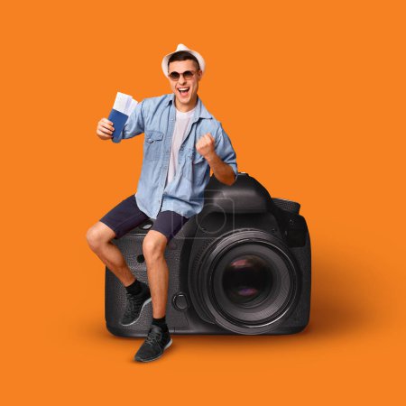 Téléchargez les photos : Joyful young european man tourist in sunglasses sit on huge photo camera, hold passport and tickets, ready to fly and trip, isolated on orange background, studio, collage, full length. Journey, travel - en image libre de droit