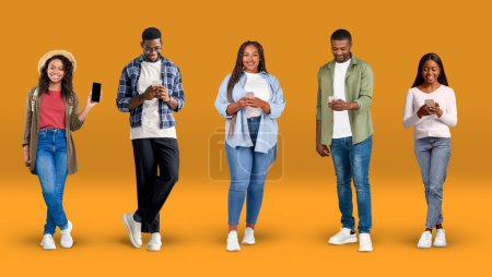 Foto de Cheerful young african american people in casual typing on phones, show phone with blank screen, on orange background, studio panorama. Communication and chat online, social media technology and app - Imagen libre de derechos