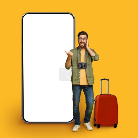 Photo for Wow, cool offer. Excited man with red suitcase traveler pointing at big blank smartphone display, showing huge white empty cellphone screen, standing over yellow studio background, mockup, collage - Royalty Free Image