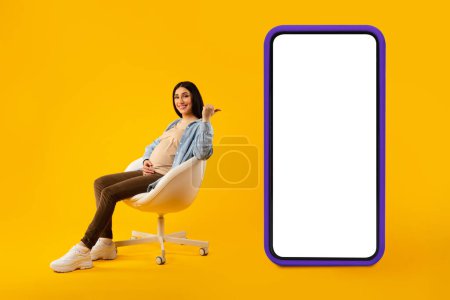 Photo for Mobile offer. Positive expectant woman pointing back at big smartphone with blank screen, sitting on chair over yellow studio background, full length shot, mockup - Royalty Free Image