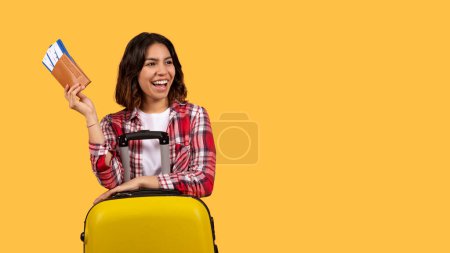 Téléchargez les photos : Happy beautiful middle eastern young lady in casual outfit tourist posing on yellow studio background, holding passport and flight tickets, standing by yellow suitcase, panorama with copy space - en image libre de droit