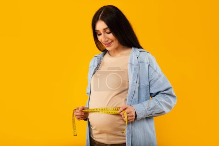 Téléchargez les photos : Positive pregnant woman measuring her belly with tape and smiling, standing on yellow background, free space. Healthy pregnancy, prenatal care, maternity, health, gynecology, medicine - en image libre de droit