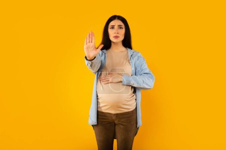 Photo for Young expectant woman doing stop sing with palm of the hand and touching belly, standing over yellow background. Warning expression with negative and serious gesture on the face - Royalty Free Image