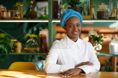 Téléchargez les photos : Portrait of pretty young black woman wearing blue turban smiling at camera while chilling at cafe, coffee shop, sitting at table alone, waiting for friends or lover, copy space for ad - en image libre de droit