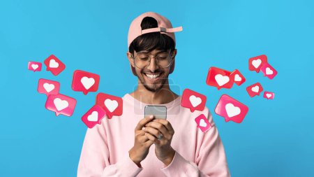 Foto de Happy young arab guy in glasses has romantic chat with hearts on phone, enjoy message on blue studio background, panorama. App for dating remotely, relationships, love at new normal, social networks - Imagen libre de derechos