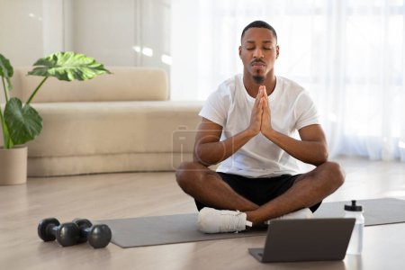 Photo for Peaceful relaxed young african american man in sportswear sitting on fitness mat in lotus pose with closed eyes, meditating at home, watching video lesson on Internet, using laptop, copy space - Royalty Free Image