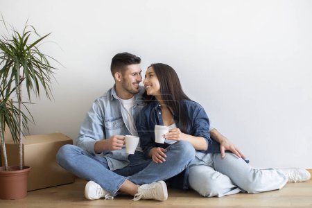 Téléchargez les photos : Portrait d'un jeune couple romantique se relaxant sur le sol et buvant du café, aimant les conjoints du millénaire assis parmi des boîtes en carton déballées, se reposant ensemble après avoir déménagé dans leur nouvelle maison, Copier l'espace - en image libre de droit