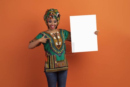 Foto de Excited happy cheerful pretty traditional african young woman pointing at empty white placard for advertisement in her hand, black lady wearing national costume, orange studio background, mockup - Imagen libre de derechos