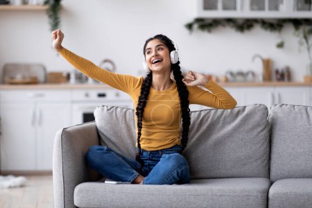 Foto de Portrait Of Cheerful Young Indian Woman Listening Music In Wireless Headphones At Home, Carefree Hindu Female Dancing And Having Fun While Sitting On Couch In Living Room, Enjoying Favorite Playlist - Imagen libre de derechos