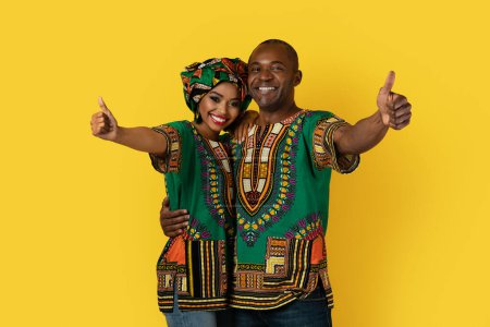 Photo for Cheery happy loving african husband and wife in bright traditional clothing embracing, showing thumb ups and smiling at camera, isolated on yellow studio background - Royalty Free Image