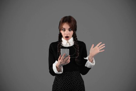 Photo for Shocked young girl student in black clothes with pigtails with open mouth reads message on smartphone, isolated on gray studio background. Victory emotions, surprise, app for study, knowledge and blog - Royalty Free Image