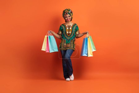 Téléchargez les photos : Mignon joyeux positif attrayant traditionnel jeune femme africaine portant un costume national lumineux et maquillage tenant des sacs à provisions colorés, isolé sur fond de studio orange, profiter du shopping - en image libre de droit