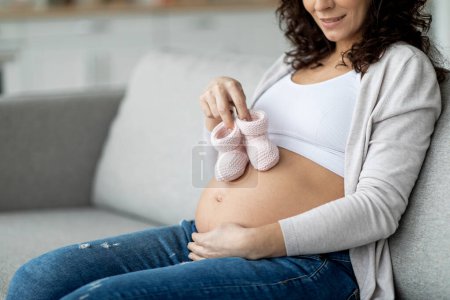 Photo for Maternity Concept. Pregnant female embracing belly and holding small baby shoes in hands while sitting on couch at home, happy expectant woman enjoying pregnancy time, closeup with copy space - Royalty Free Image