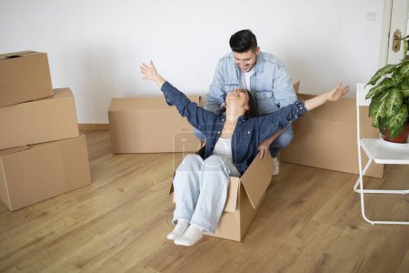 Photo for Young Man Riding His Happy Wife In Cardboard Box On Moving Day, Cheerful Young Couple Fooling In Their New Home, Happy Spouses Having Fun While Unpacking Things During Relocation, Copy Space - Royalty Free Image