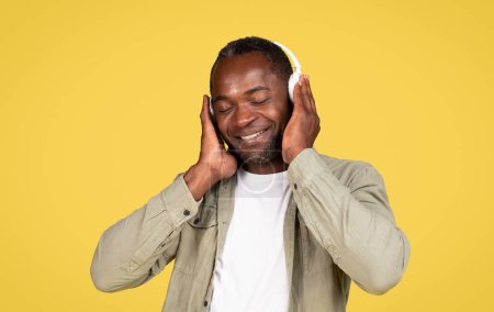 Téléchargez les photos : Smiling relaxed adult black guy in casual, wireless headphones enjoying music in free time, isolated on yellow background, studio. Listen song and audio app, ad and offer, fun and entertainment alone - en image libre de droit