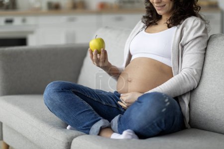 Téléchargez les photos : Jeune femme enceinte tenant une pomme verte biologique et caressant le ventre tout en étant assise sur le canapé à la maison, méconnaissable s'attendant à ce que la femme jouisse d'une alimentation saine pendant la grossesse, image recadrée - en image libre de droit