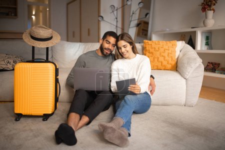 Photo for Happy young arabic man hug european lady use tablet and laptop, choose country for vacation travel in living room interior with suitcase and hat. Love, relationship, app for ticket trip and tourists - Royalty Free Image