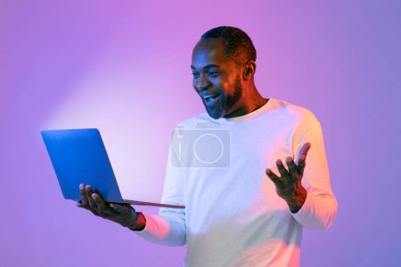 Téléchargez les photos : Incroyable bel homme afro-américain d'âge moyen en blanc en utilisant un tout nouveau portable, en regardant l'écran brillant de l'ordinateur portable et en gestualisant, en utilisant la plus récente application, fond de studio néon, espace de copie - en image libre de droit