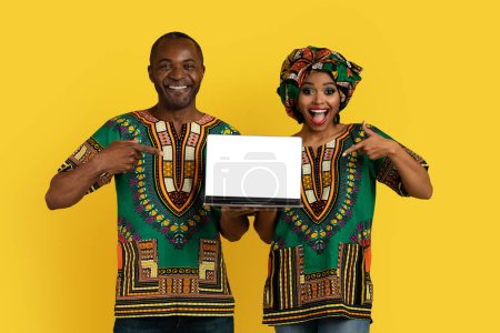 Photo for Positive happy bright loving black couple in traditional african costumes pointing at laptop white blank screen and smiling, showing online offer, yellow background, mockup - Royalty Free Image