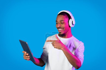 Photo for Entertainment and gadgets. Cool cheerful handsome young black guy in white pointing at brand new pc digital pad at his hand and smiling, using wireless headset on blue in neon light, copy space - Royalty Free Image